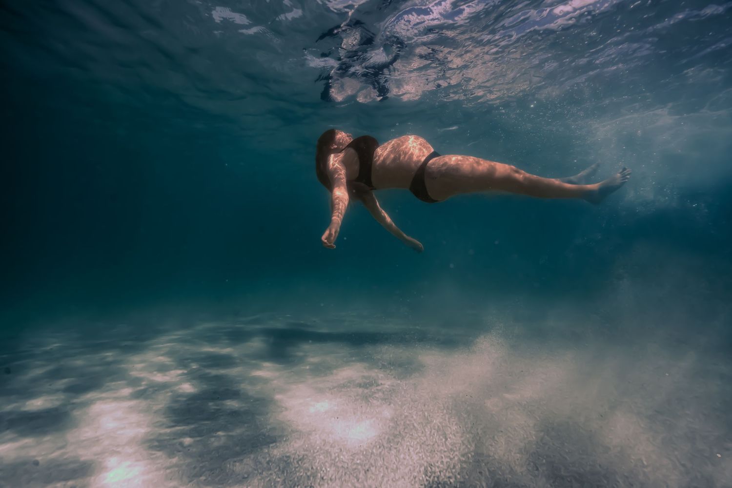 A person swims underwater in a deep blue ocean with sunlight streaming through the surface creating ethereal rays.