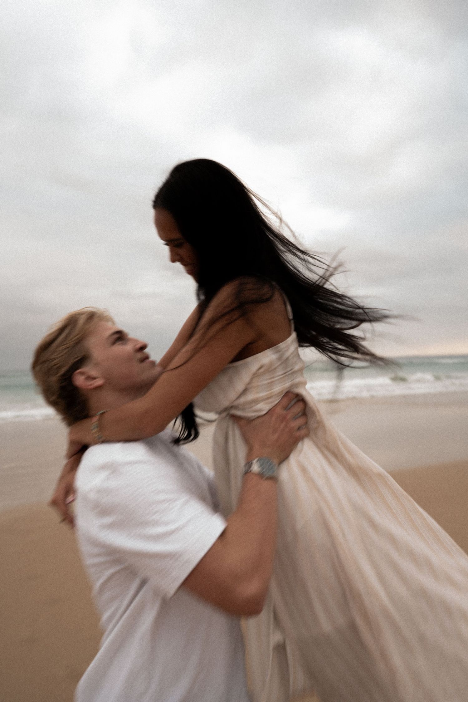 A romantic couple shares an intimate moment on a cloudy beach as the wind blows through their hair.