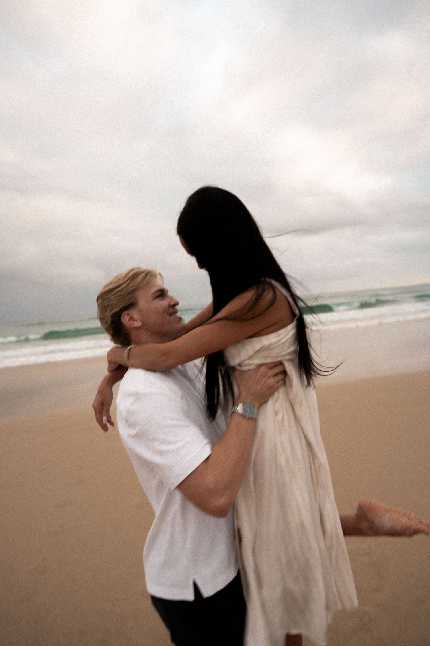 Two people in casual attire playfully interacting on a cloudy beach with ocean waves in the background.