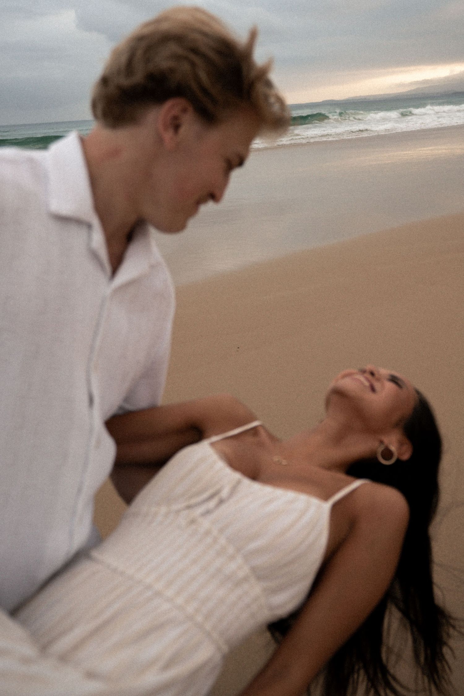 A couple in white clothing embrace tenderly on a sandy beach with ocean waves in the background.