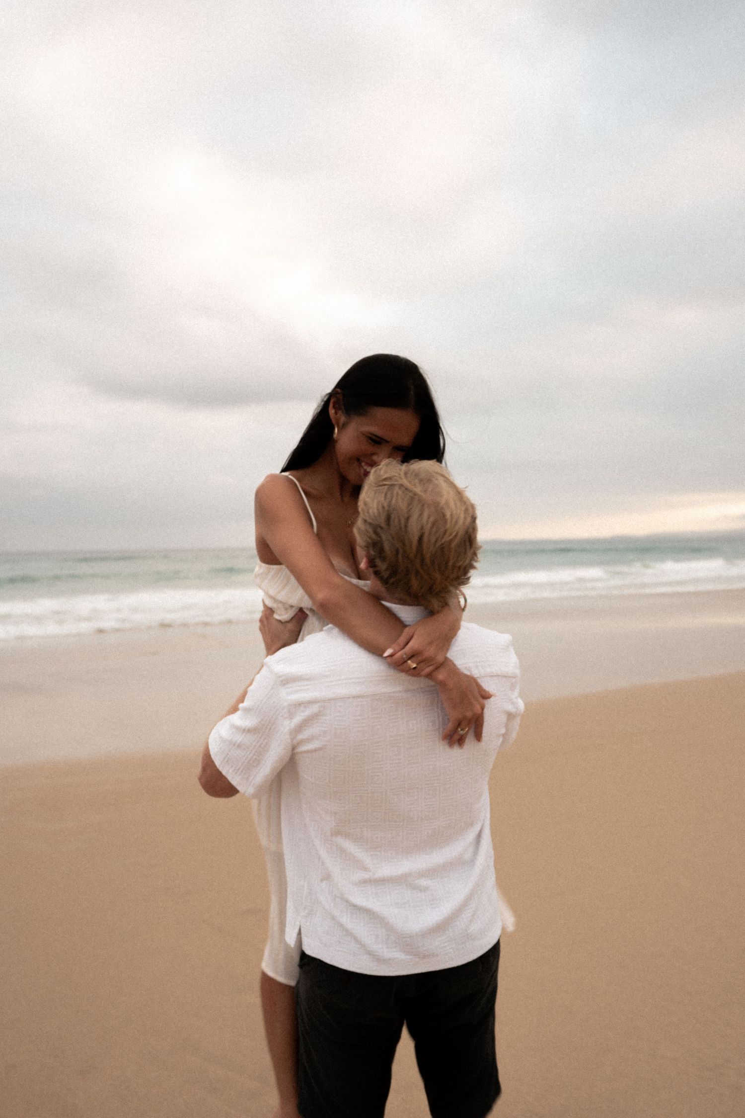 A tender embrace between two people wearing white shirts on a misty beach at sunset.