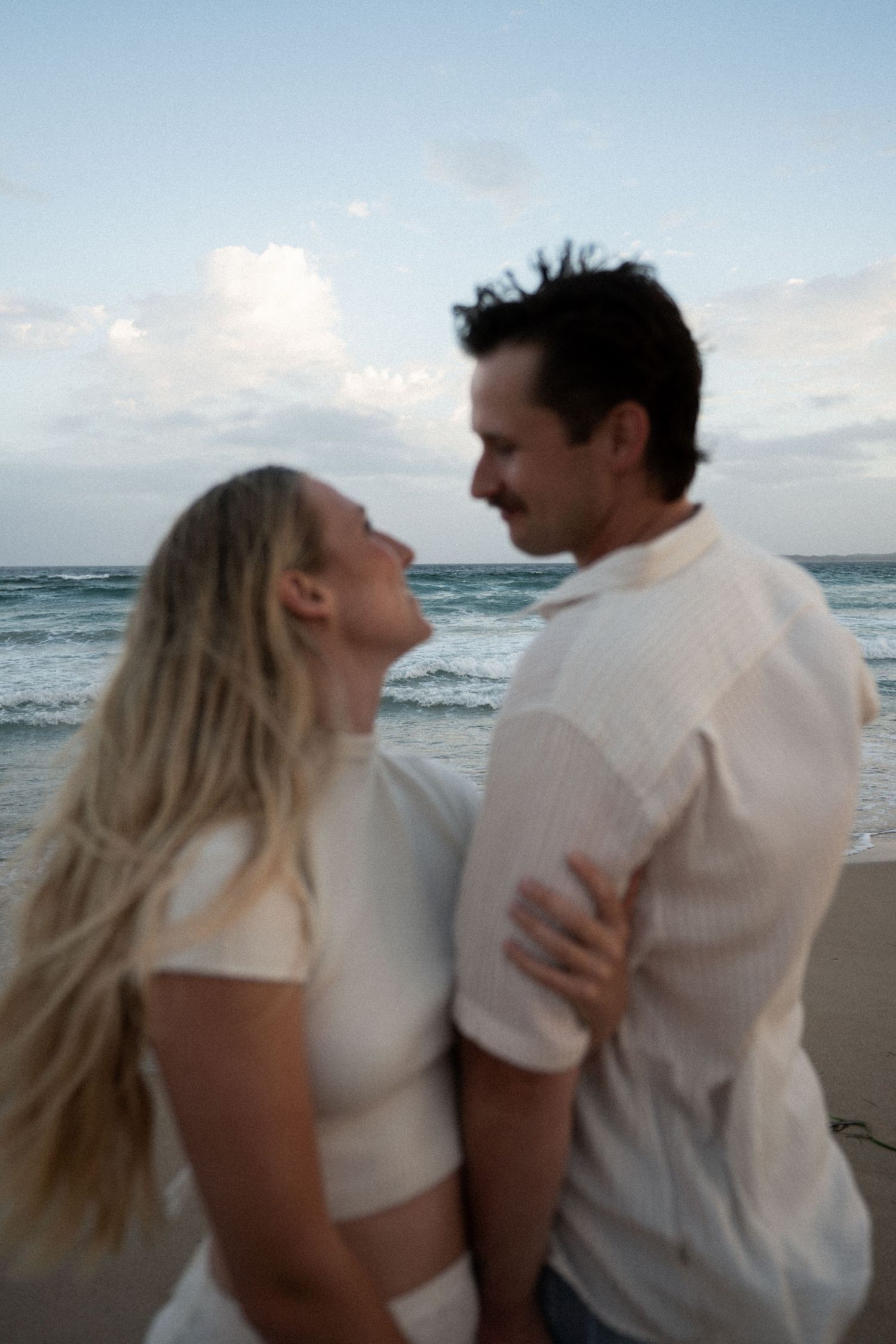 A couple in white outfits shares a romantic moment on a beach at sunset with waves crashing behind them.
