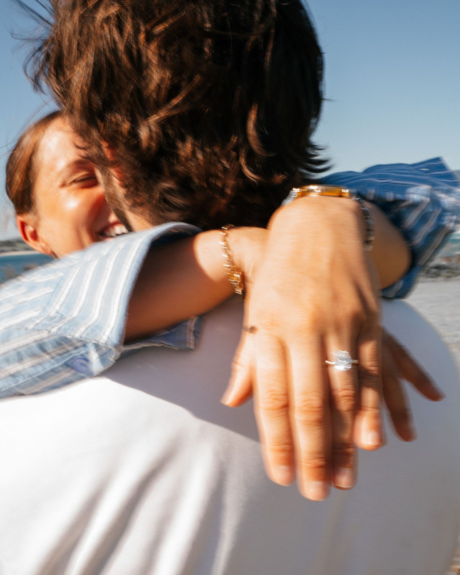Close-up of two people embracing while showing off rings and jewelry on their hands in sunlight.