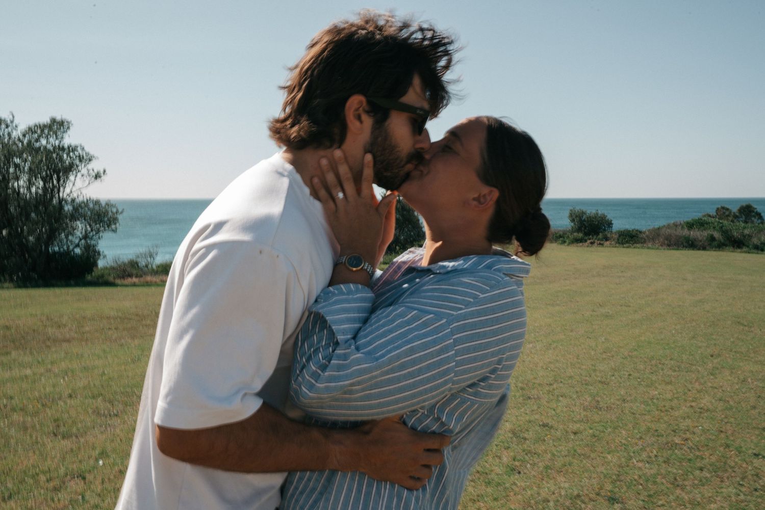 A couple shares a romantic kiss in a scenic grassy field overlooking the ocean on a sunny day.