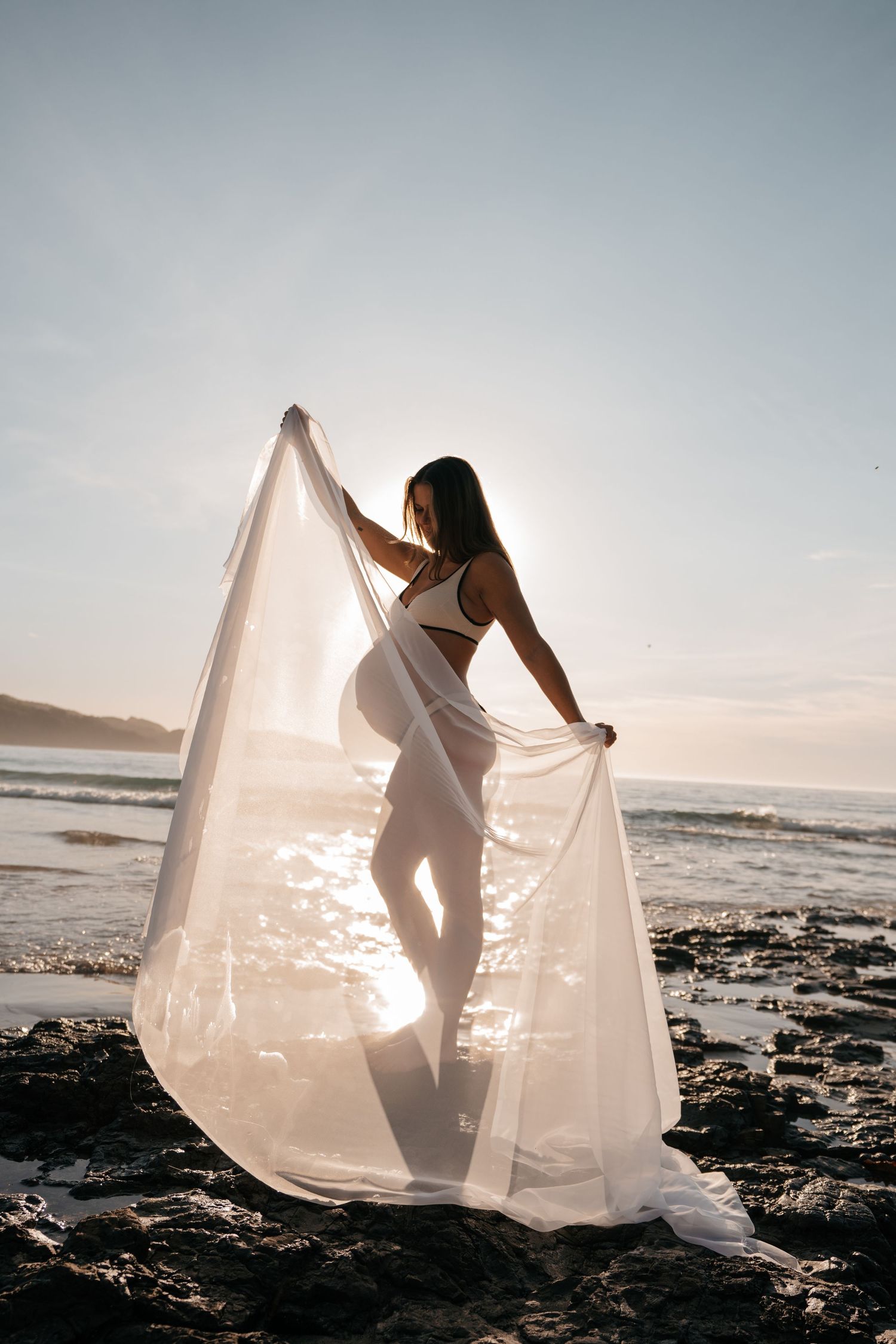 A silhouetted figure dances with flowing white fabric on a rocky beach at sunset, creating an ethereal scene.