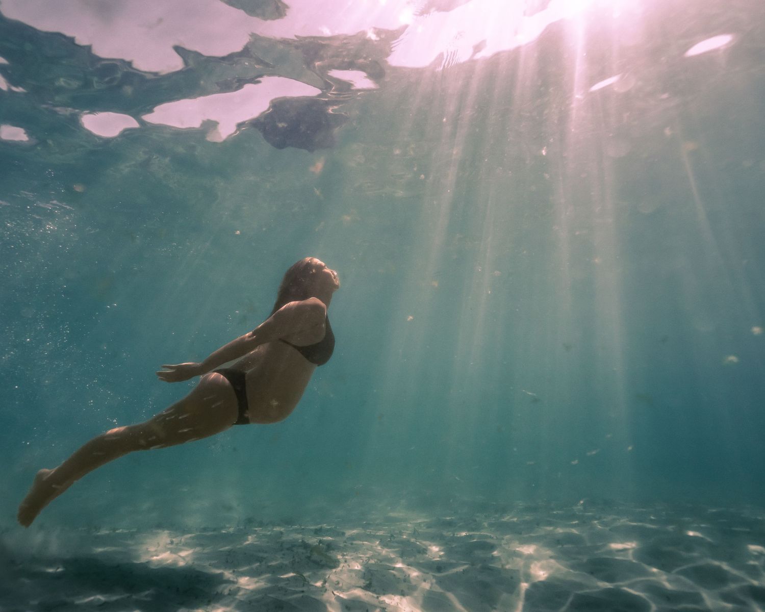 Underwater view of a person floating beneath sunbeams streaming through turquoise waters.