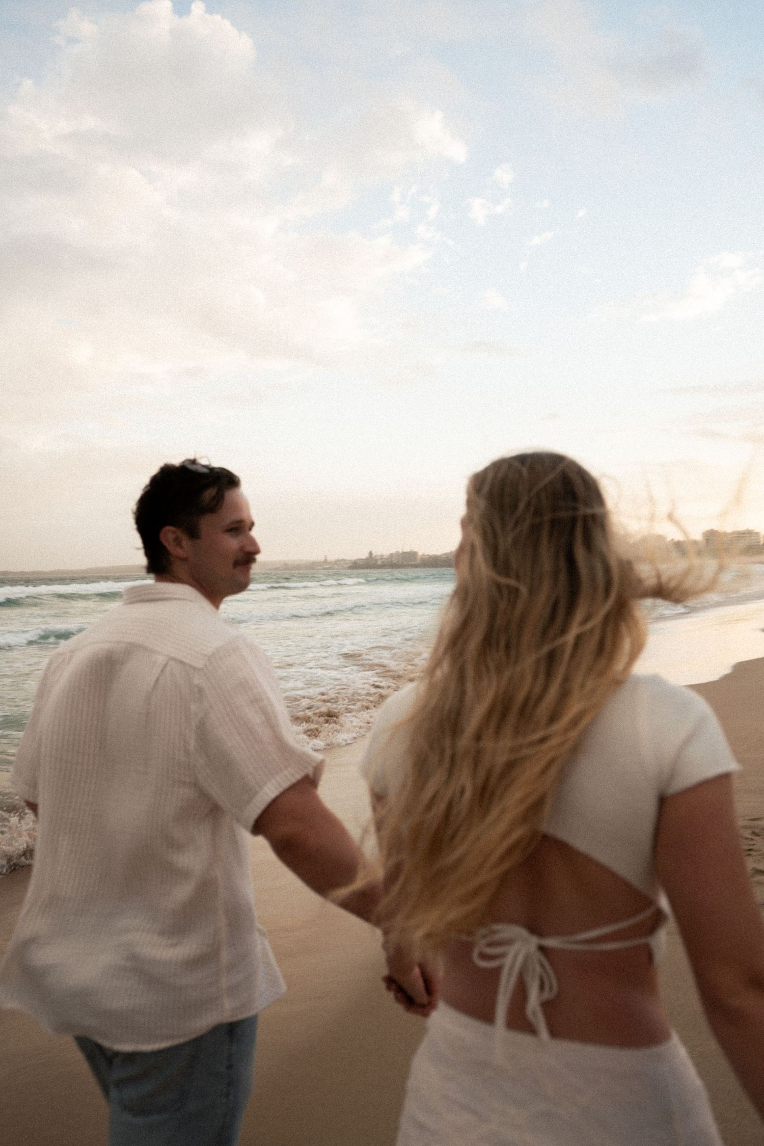 A couple walks hand in hand along a beach at sunset wearing white outfits with ocean waves in the background.