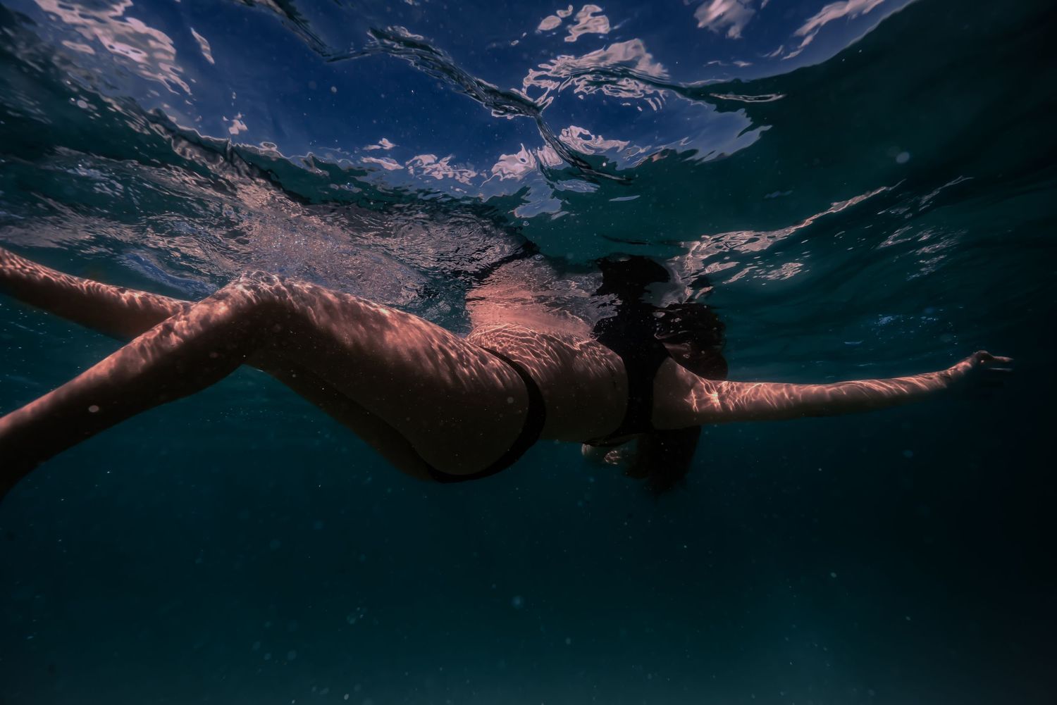 A person swims underwater in a dark pool with sunlight filtering through the surface creating a moody atmosphere.