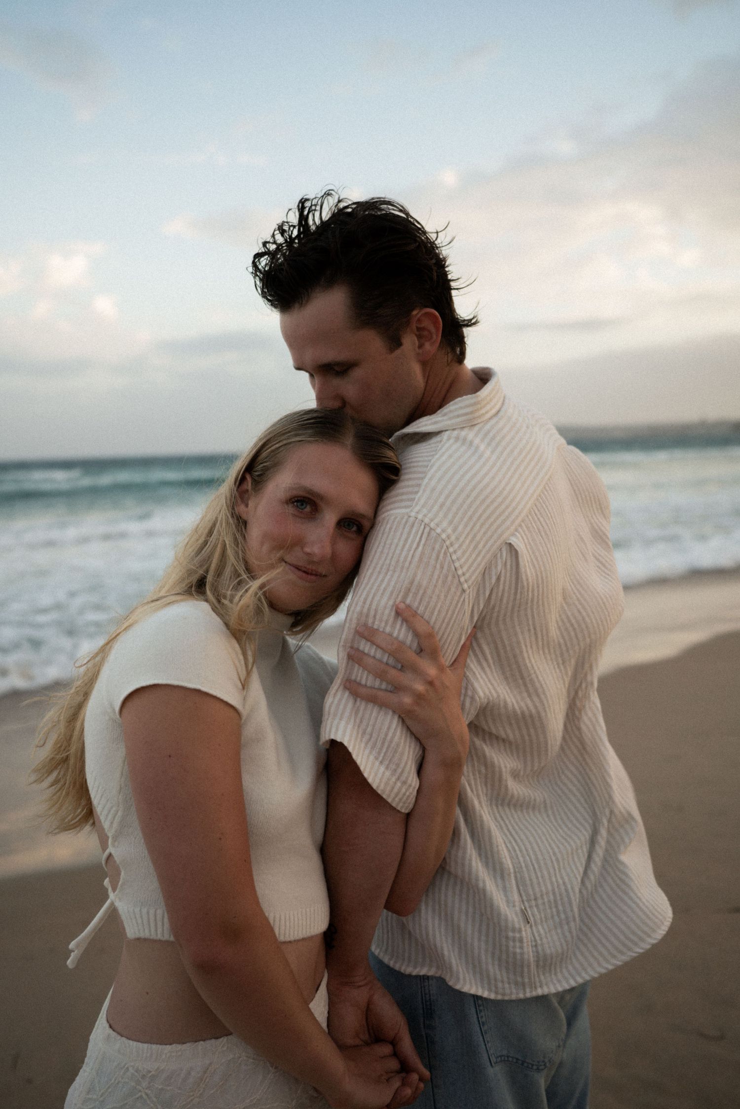 A romantic couple embraces on a beach at sunset with waves and cloudy skies in the background.