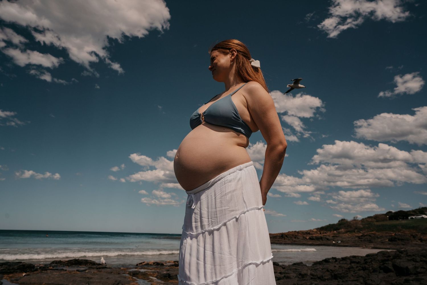 A pregnant woman in a white dress stands on a beach under a cloudy blue sky, looking towards the horizon.