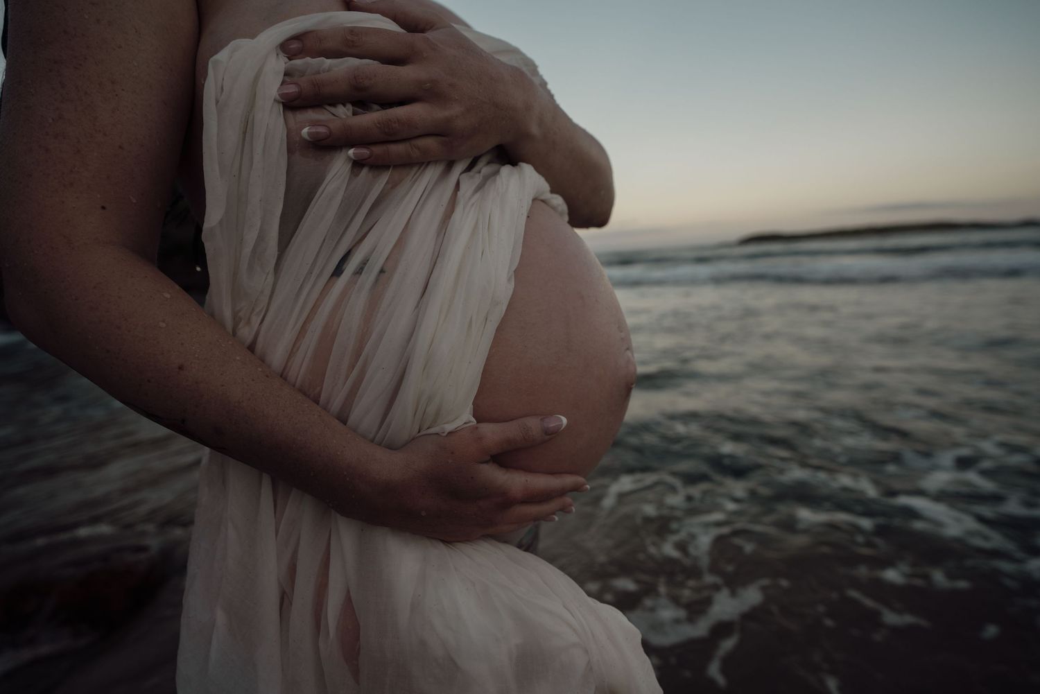 Maternity photoshoot at sunset showing hands cradling pregnant belly by the ocean.