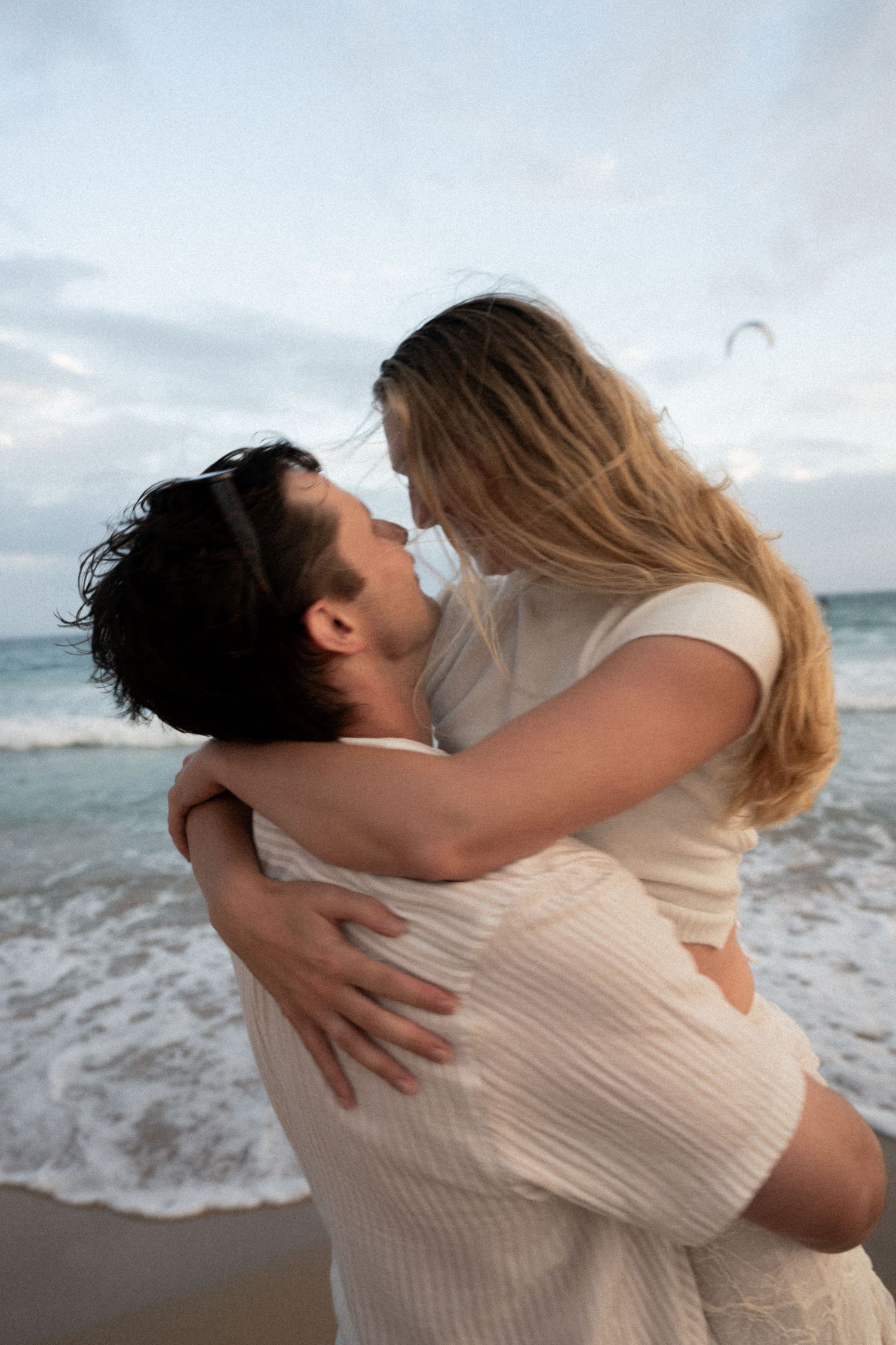 A couple embraces and spins joyfully on a beach with ocean waves in the background.