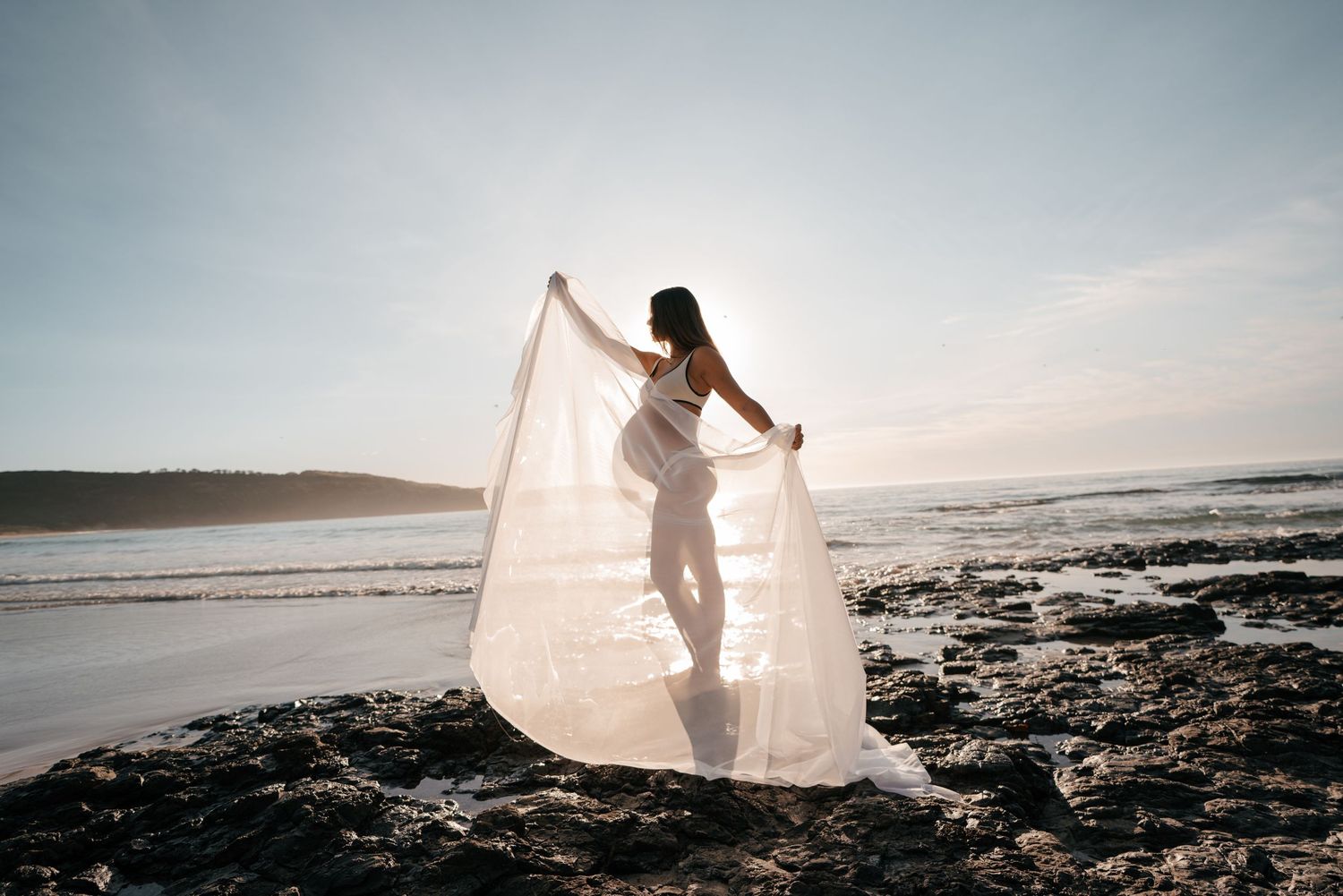 A person in white dress stands on rocky beach at sunset holding flowing fabric in dramatic coastal scene.