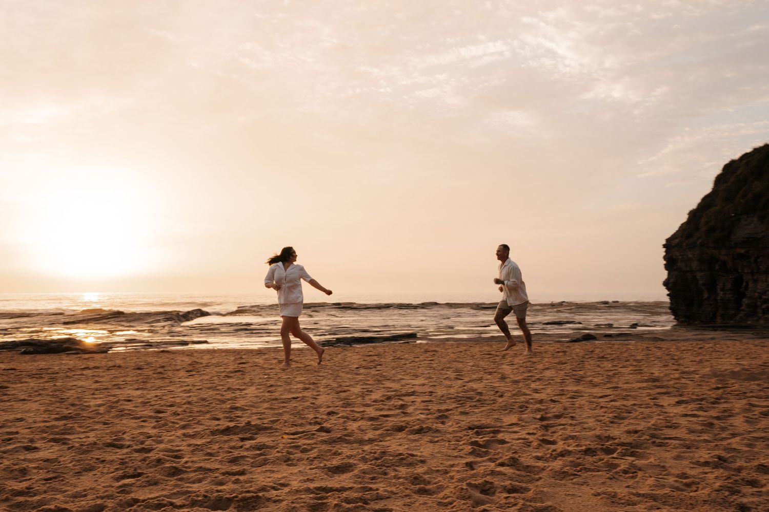 Silhouetted figures play and run along a sandy beach at sunset with dramatic orange sky and crashing waves.
