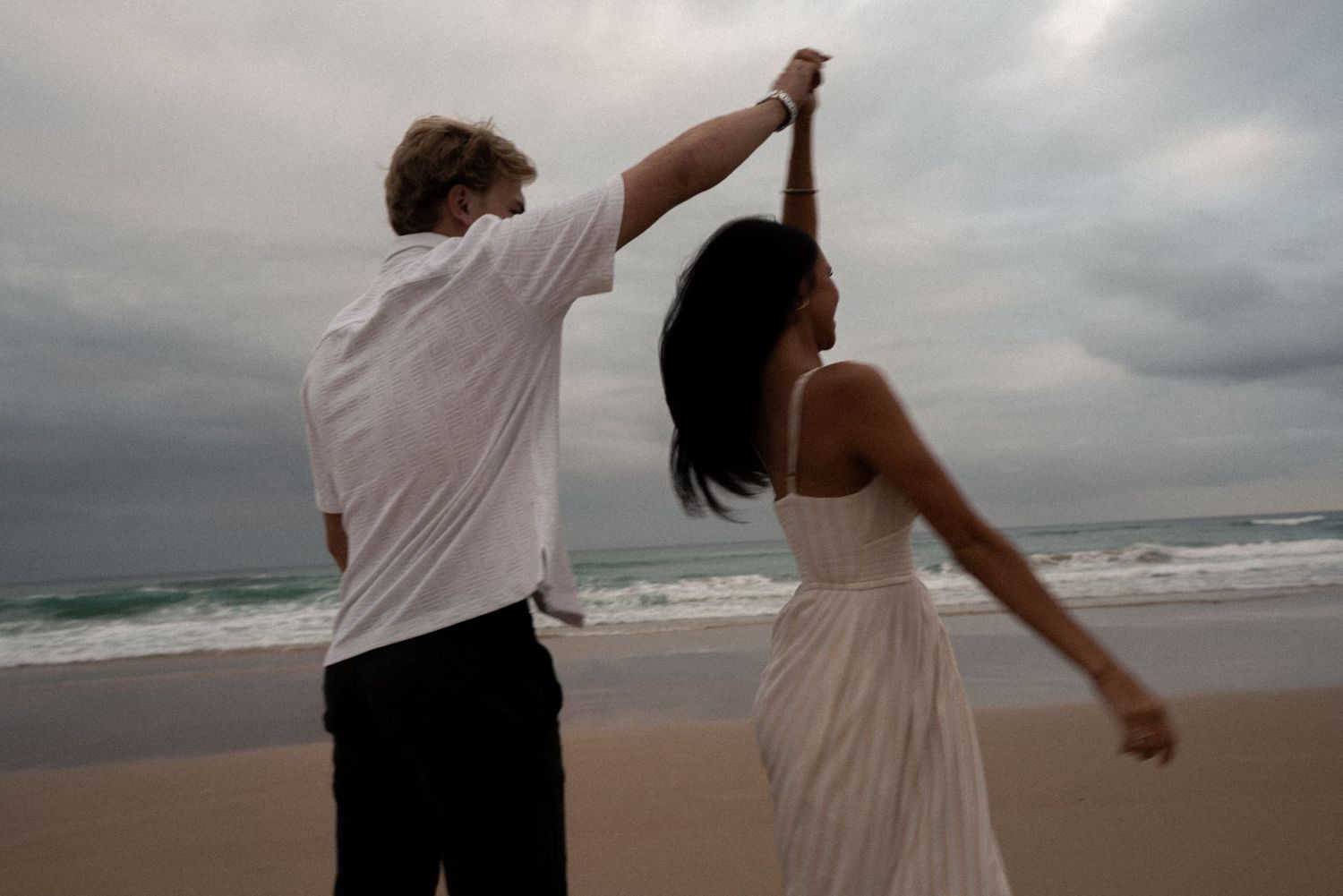 A couple in white clothing dances together on a cloudy beach at dusk.