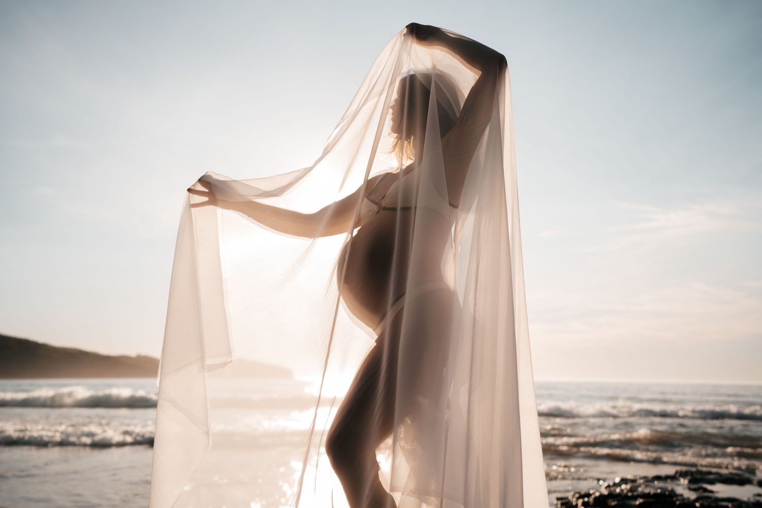 Ethereal figure draped in flowing white fabric dances gracefully along a misty beach at sunset.