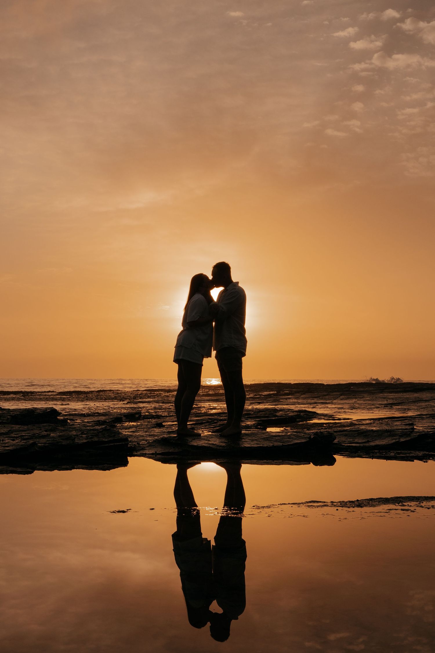 Silhouettes of a romantic couple kissing on a beach at sunset with golden sky and reflective water.