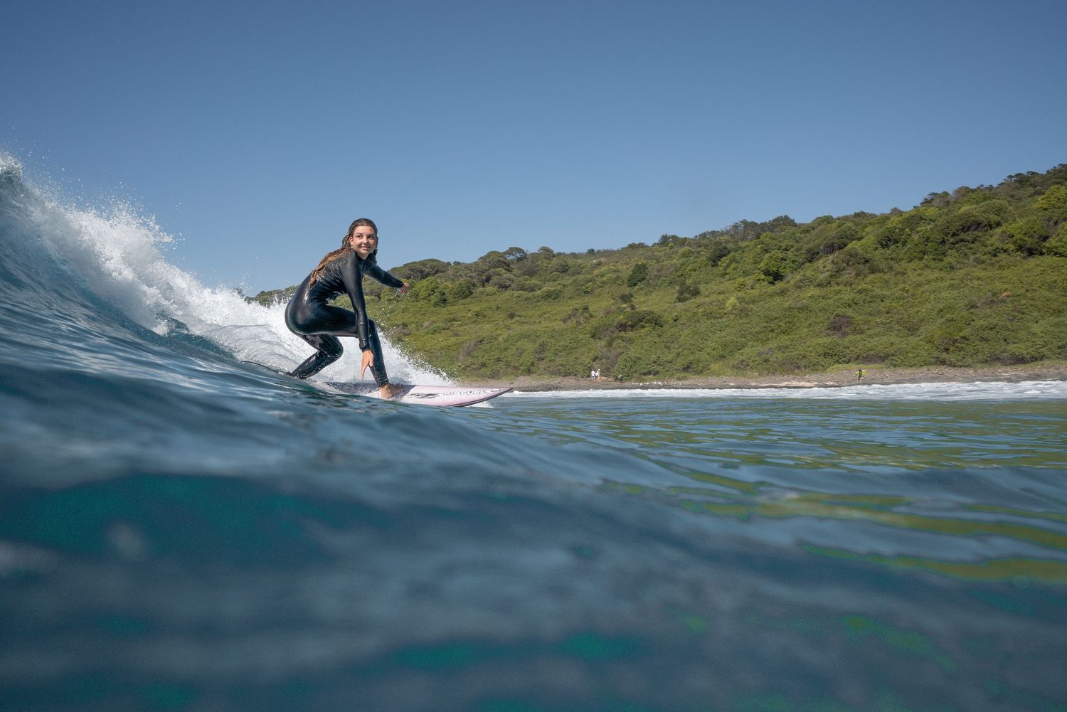 Surfer rides a wave on a sunny day at a scenic coastal location with green hills in the background.