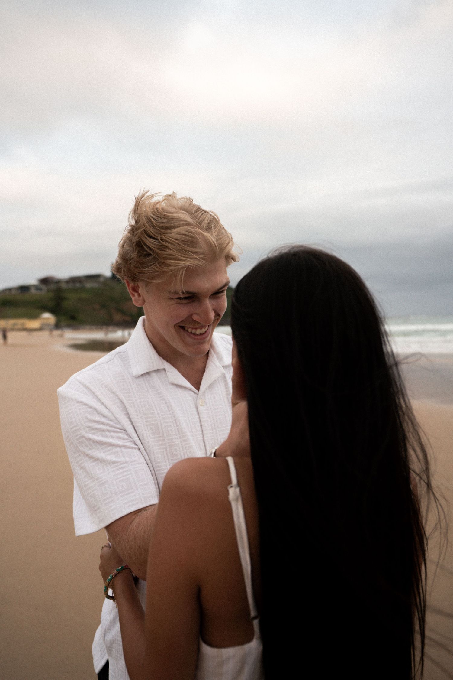 A couple shares an intimate moment together on a cloudy beach with waves in the background.