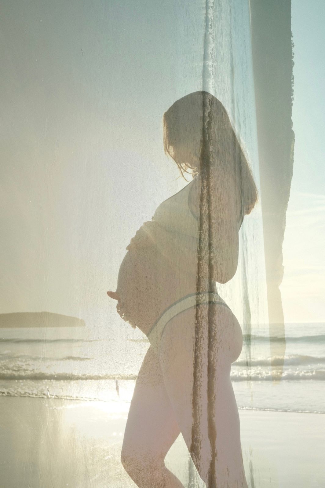 Artistic silhouette of an expectant mother standing against soft beach light with a dreamy double exposure effect.