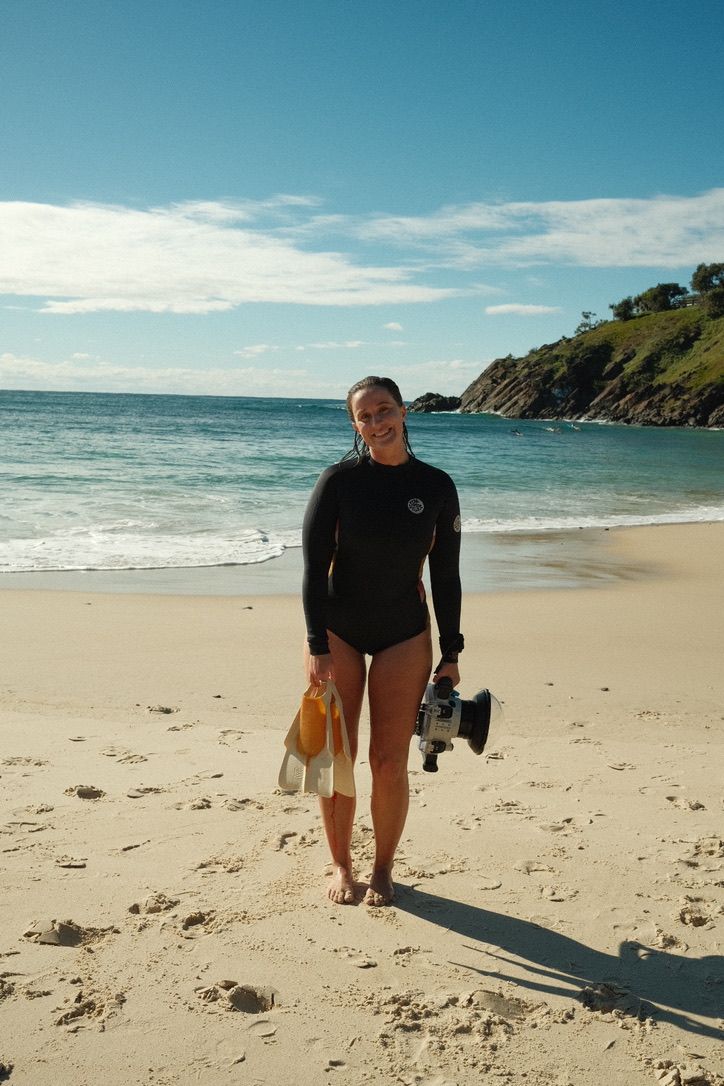 Person walking along sunny beach with camera gear and ocean cliffs in background.