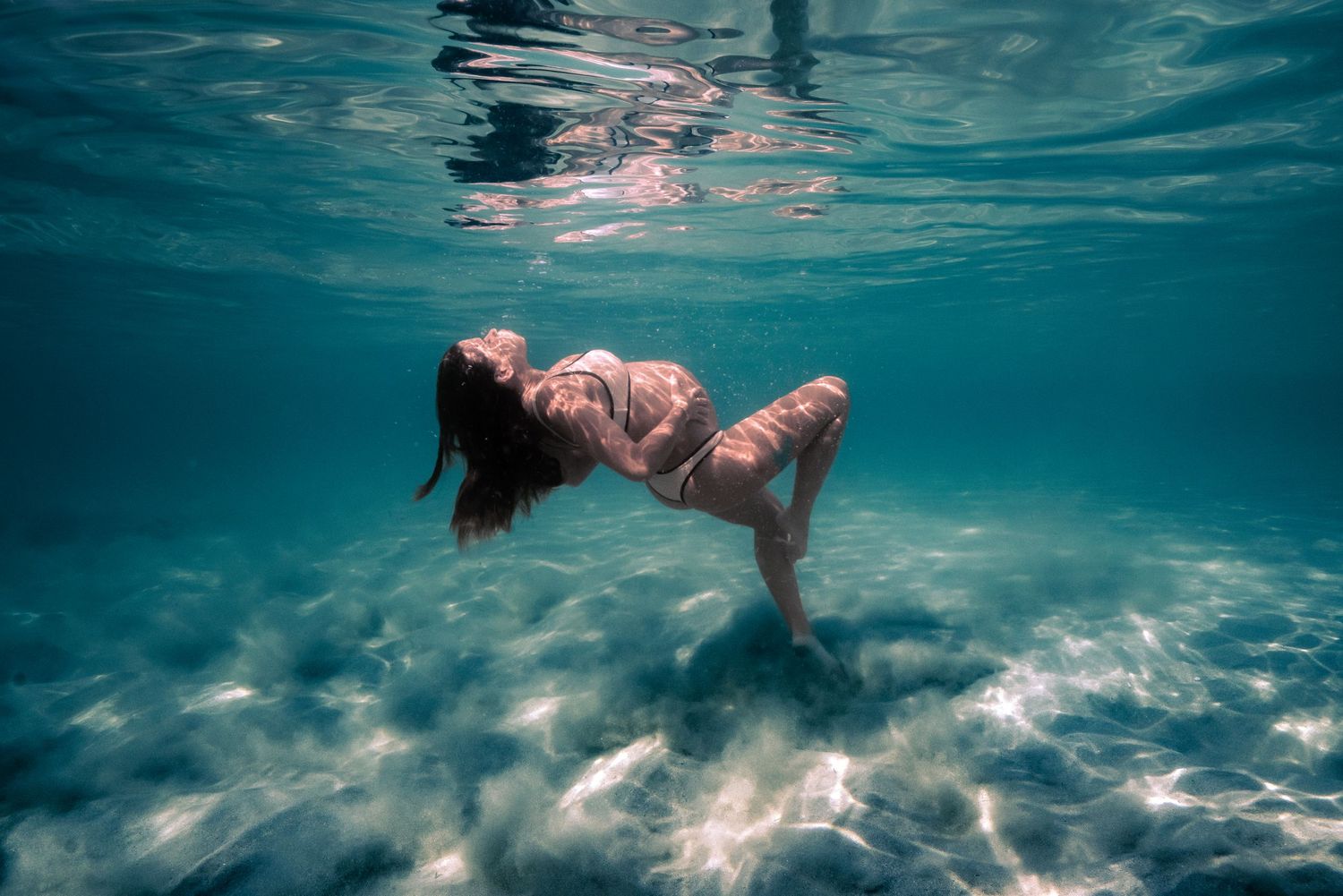 Figure floats gracefully underwater in a turquoise pool with streaming sunlight creating ethereal patterns.