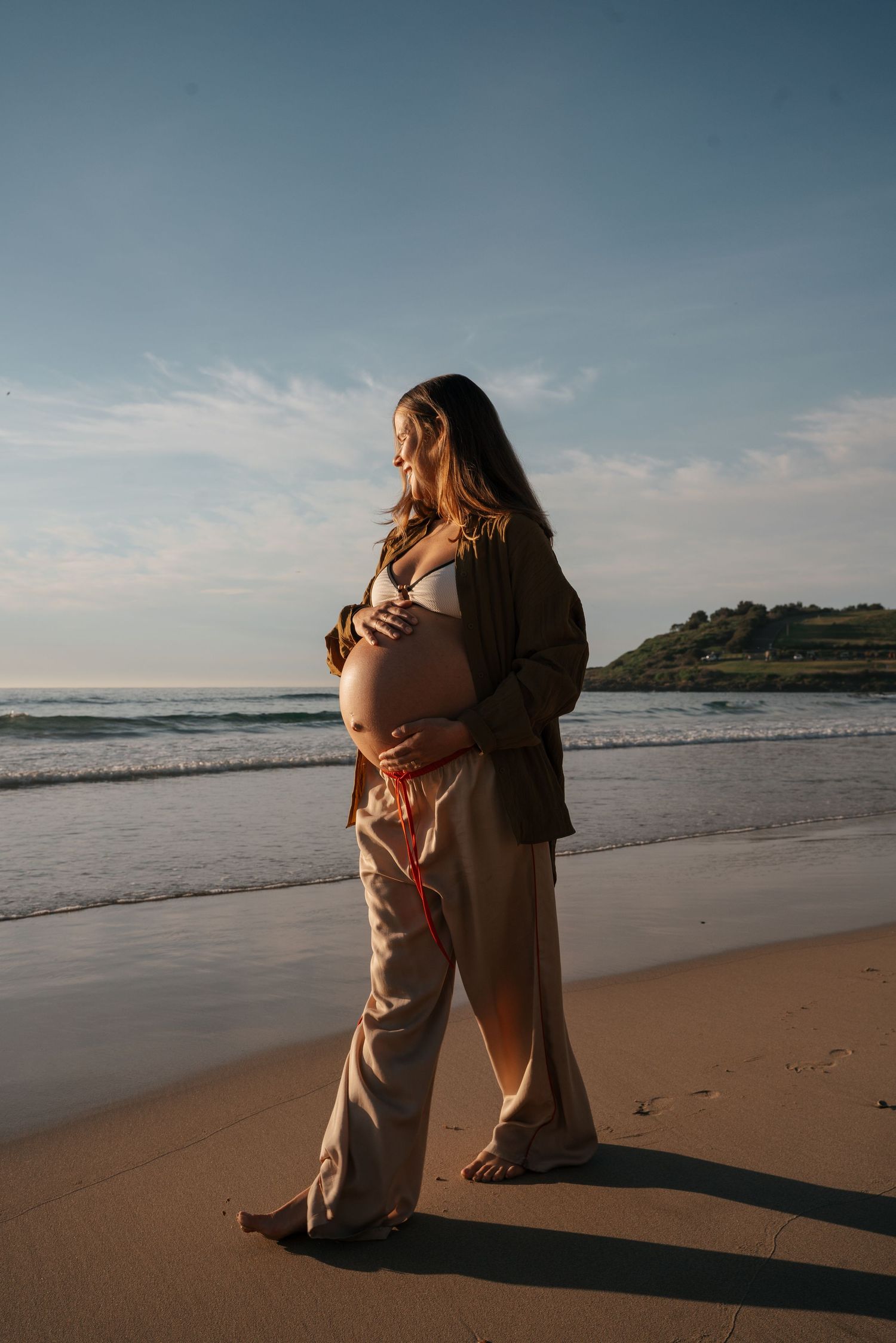 A pregnant figure stands on a peaceful beach at sunset wearing neutral-toned clothing and gazing at the ocean.