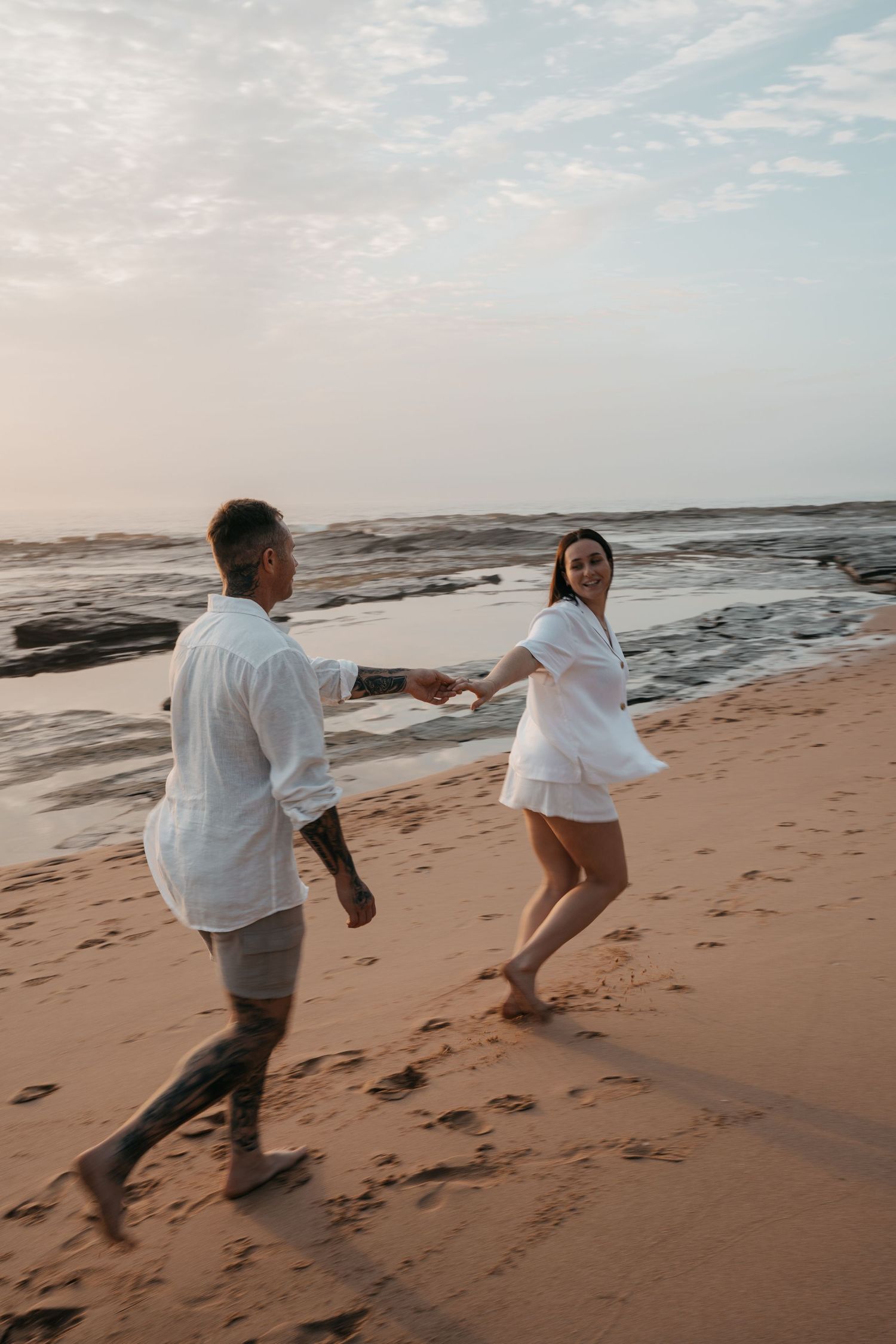 A couple in white clothing runs playfully along a sandy beach at sunset with ocean waves in the background.