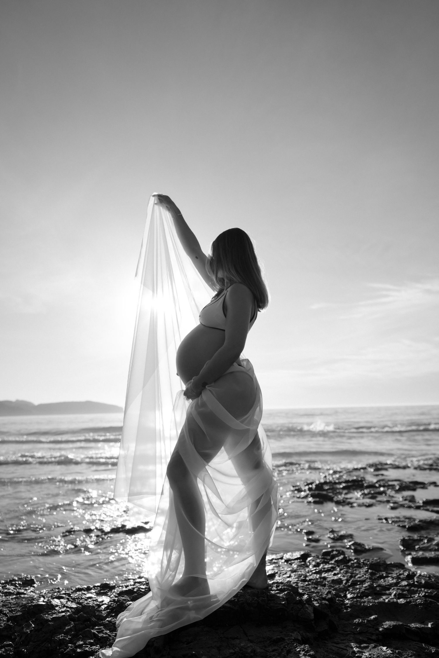 A romantic black and white photo series of a couple embracing on a rocky beach with flowing white fabric in the wind.