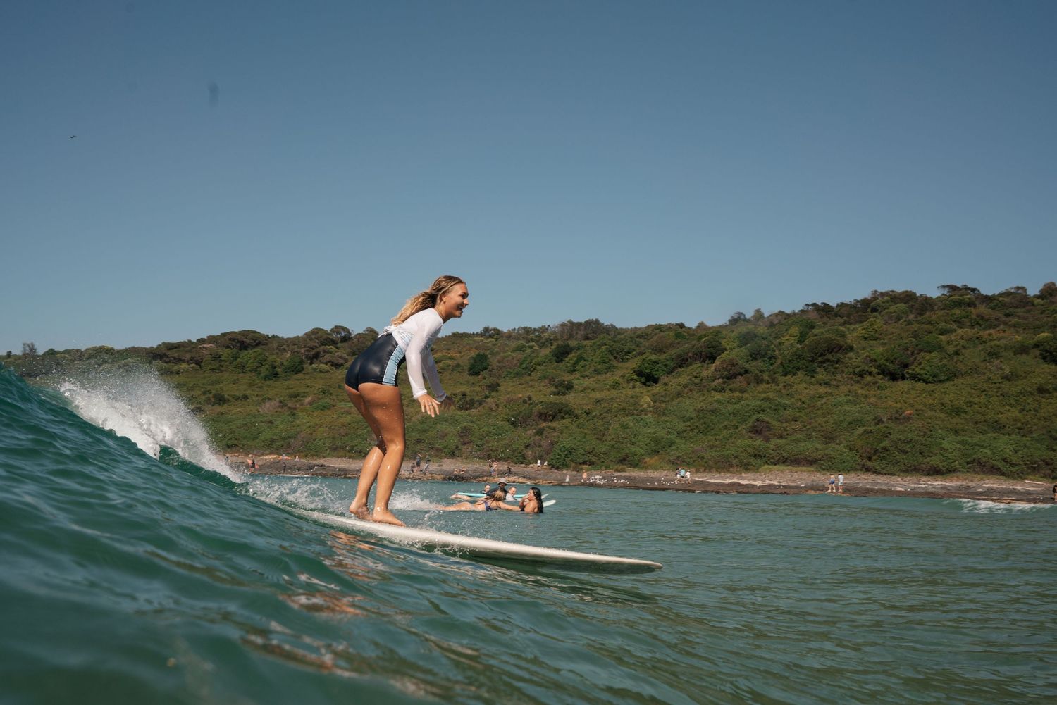 Surfer rides a longboard on a gentle wave along a scenic coastline with green hills in the background.