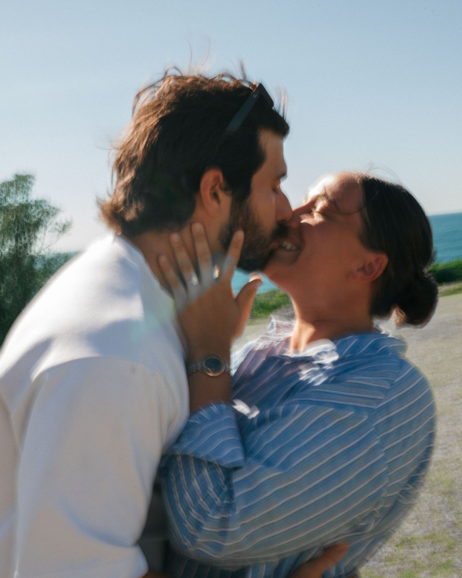 Two people in white and blue clothing share an intimate moment at the beach on a sunny day.