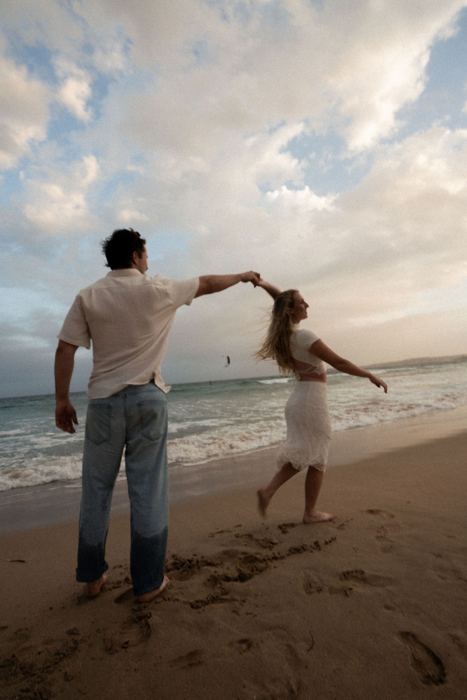 Couple playfully dancing and spinning together on a cloudy beach at sunset.