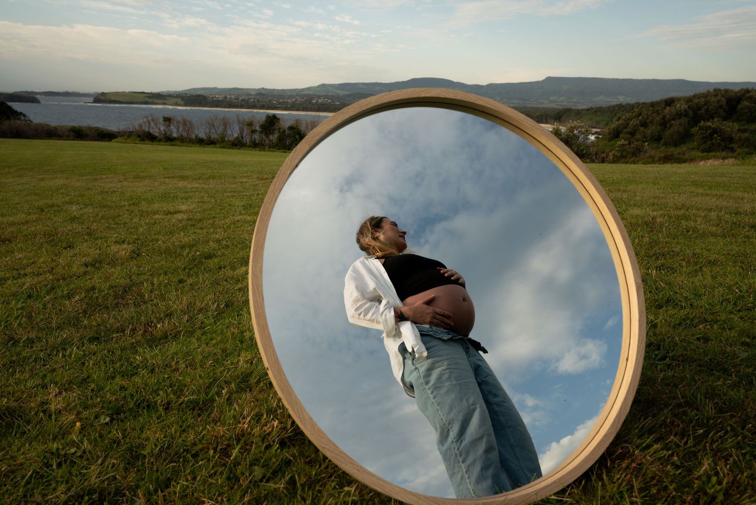 Creative maternity photos using round mirrors reflecting sky in grassy field.