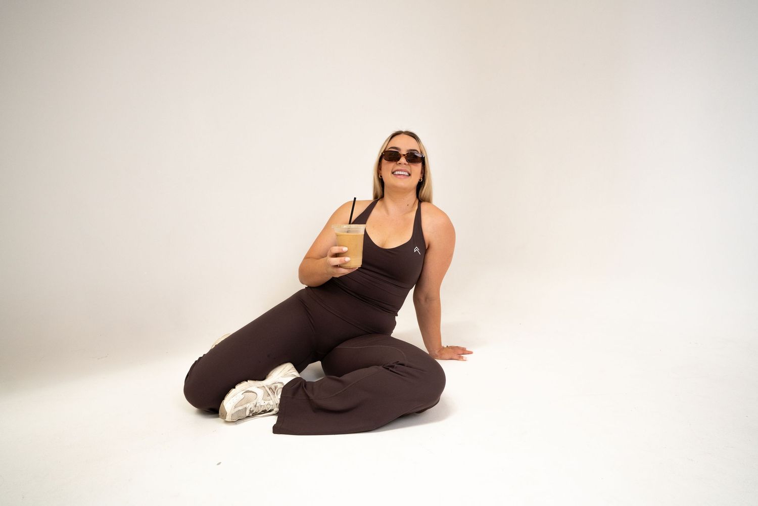 A person in black athletic wear and sunglasses sits on the floor giving a thumbs up against a white studio backdrop.