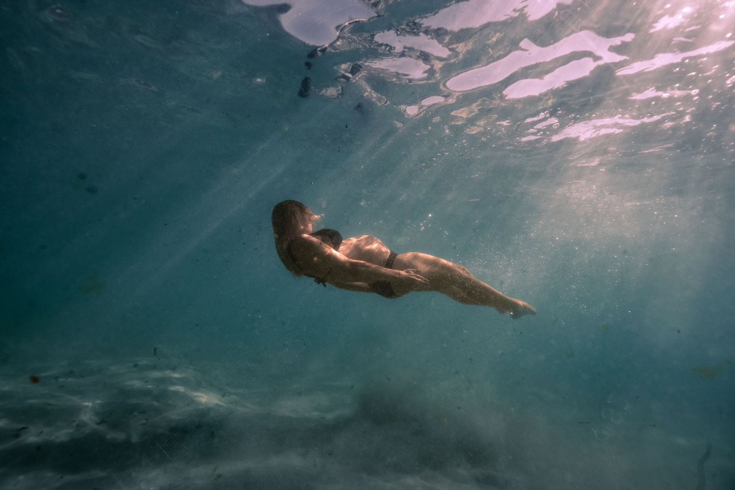 A person swimming underwater in a pool with sunlight streaming through the surface creating a dreamy atmosphere.