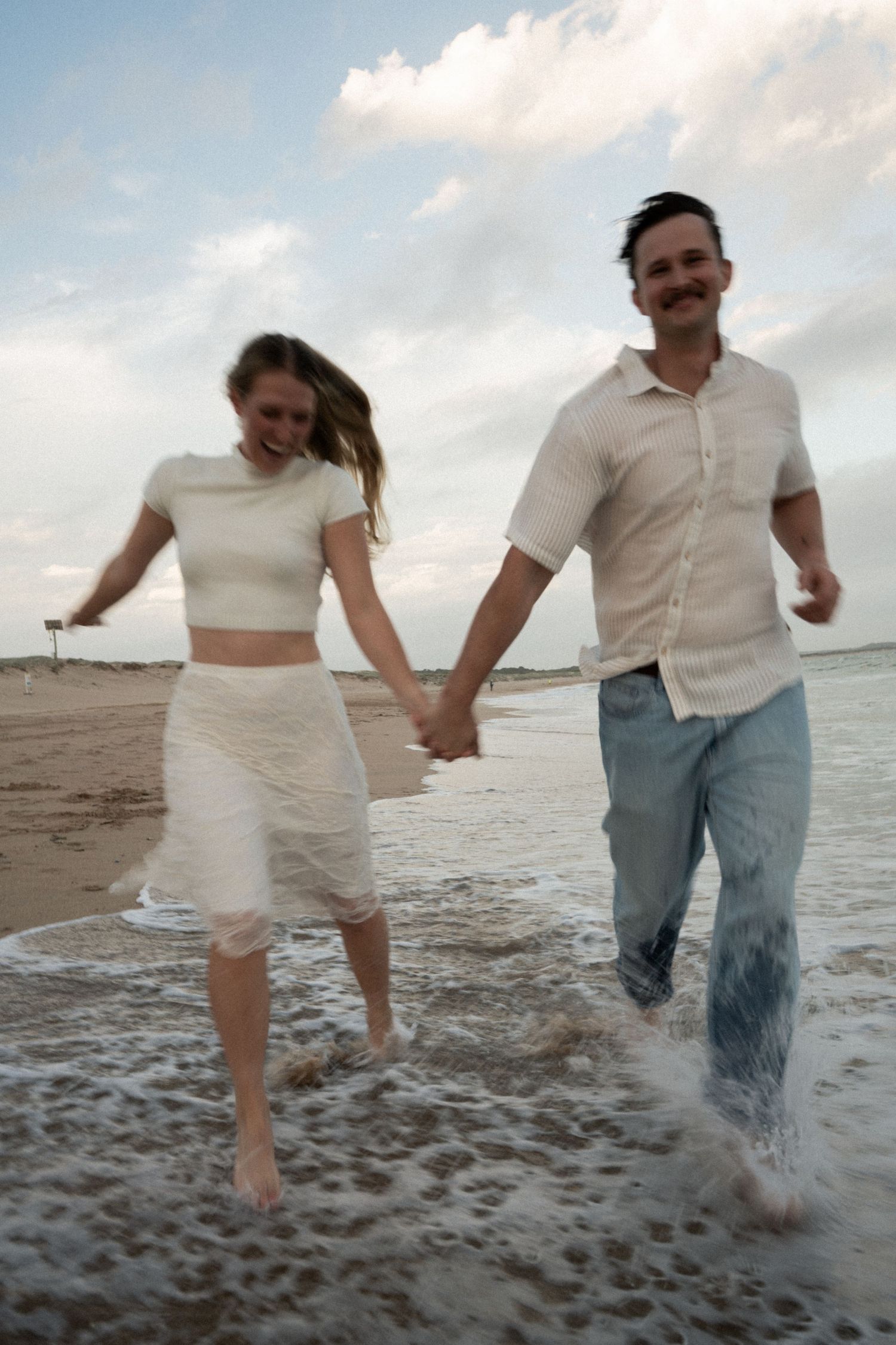 A couple runs and plays together on a sandy beach at sunset, kicking up sand as they move along the shoreline.