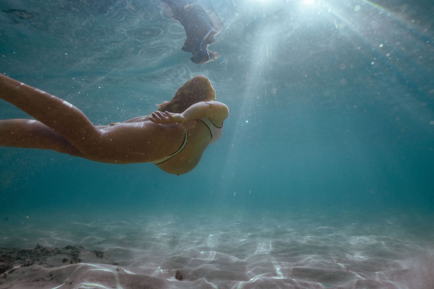 Underwater view of feet swimming in clear turquoise water with sunlight streaming through and sandy ocean floor below.