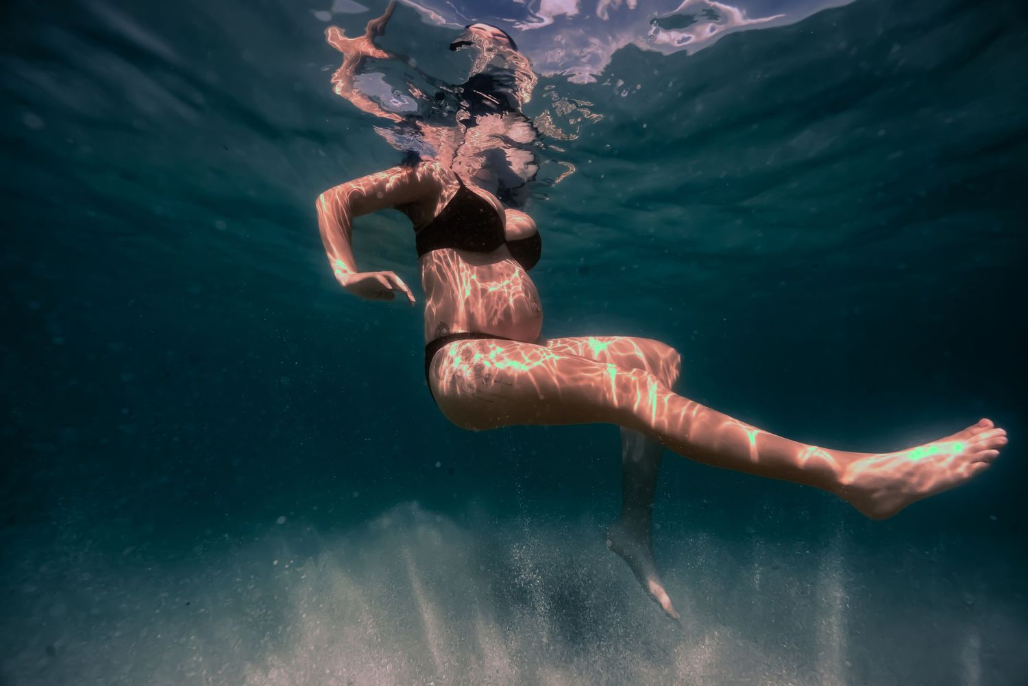 Underwater view of a person swimming in dark blue water with sunlight streaming down through the surface.