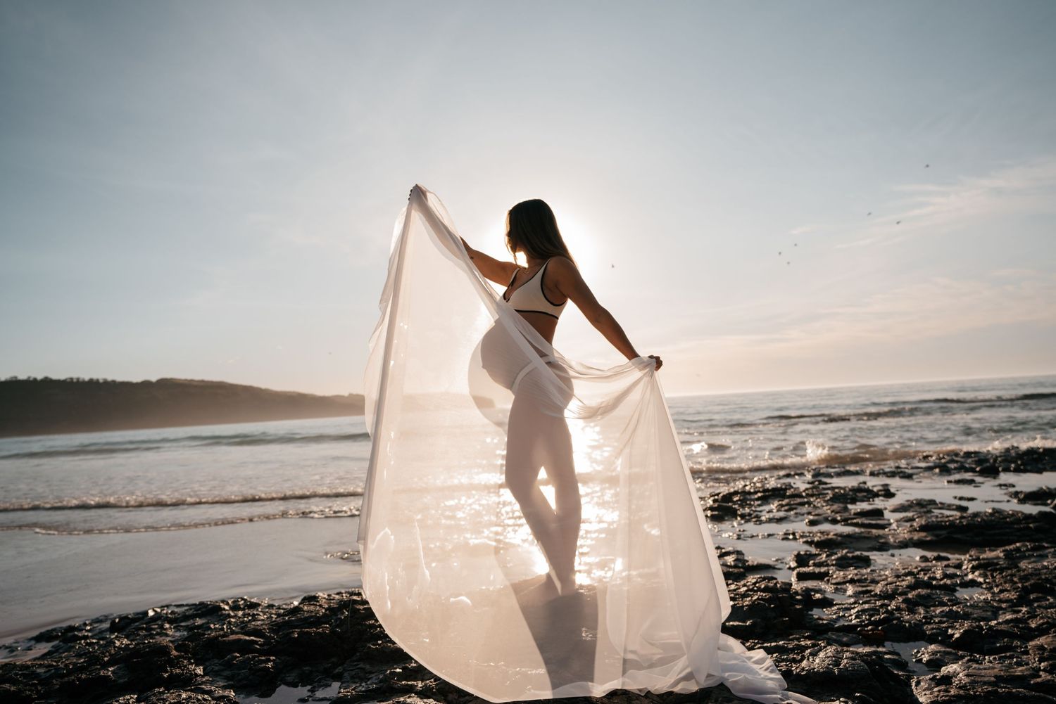 Ethereal silhouette dances with flowing white fabric on rocky beach at sunset creating dreamy coastal scene.