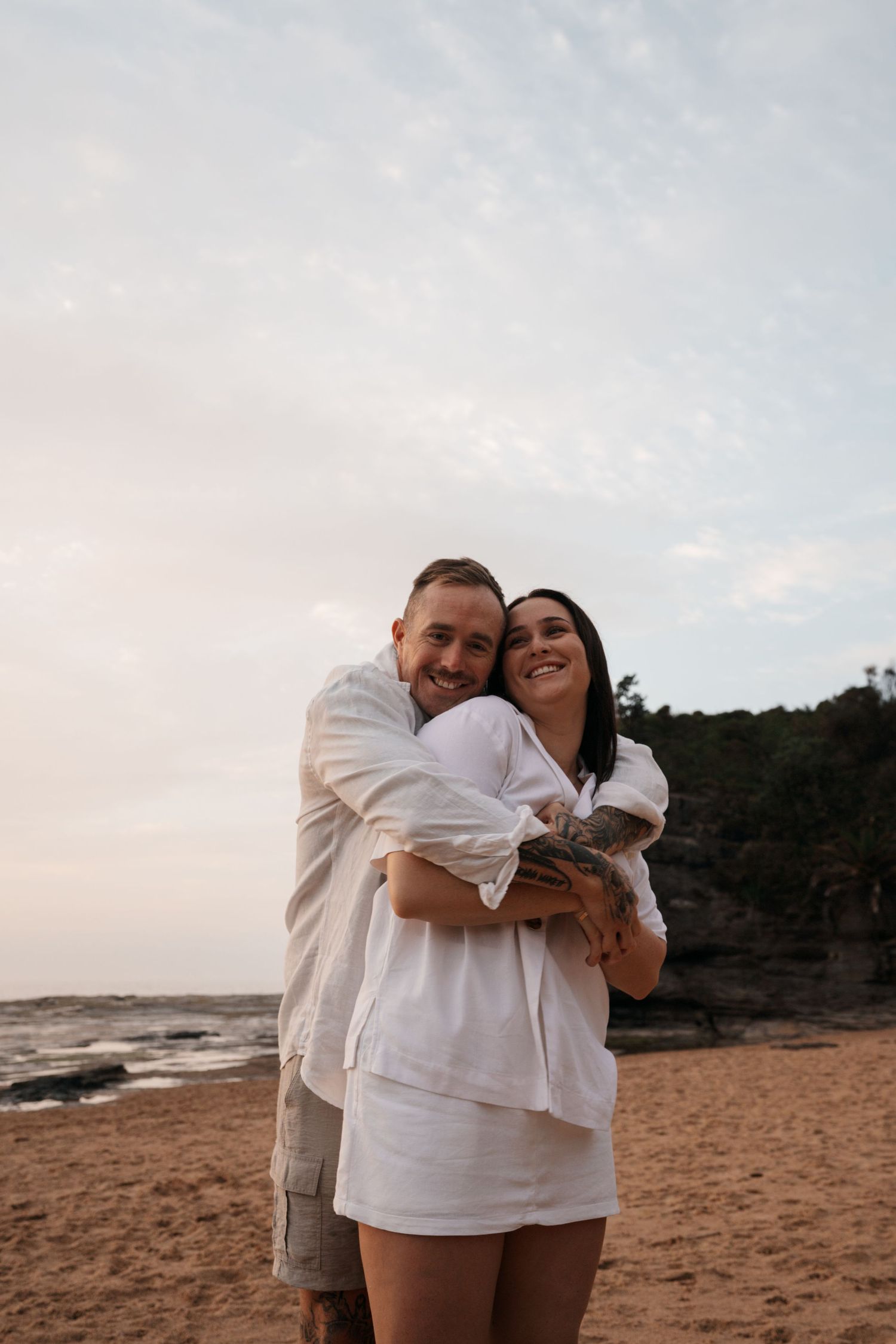 A couple embraces on a sandy beach at sunset with rocky cliffs in the background.