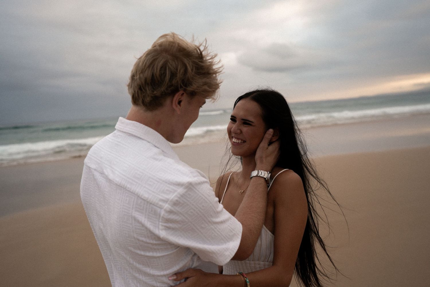 A happy couple embraces on a cloudy beach at sunset with waves crashing in the background.