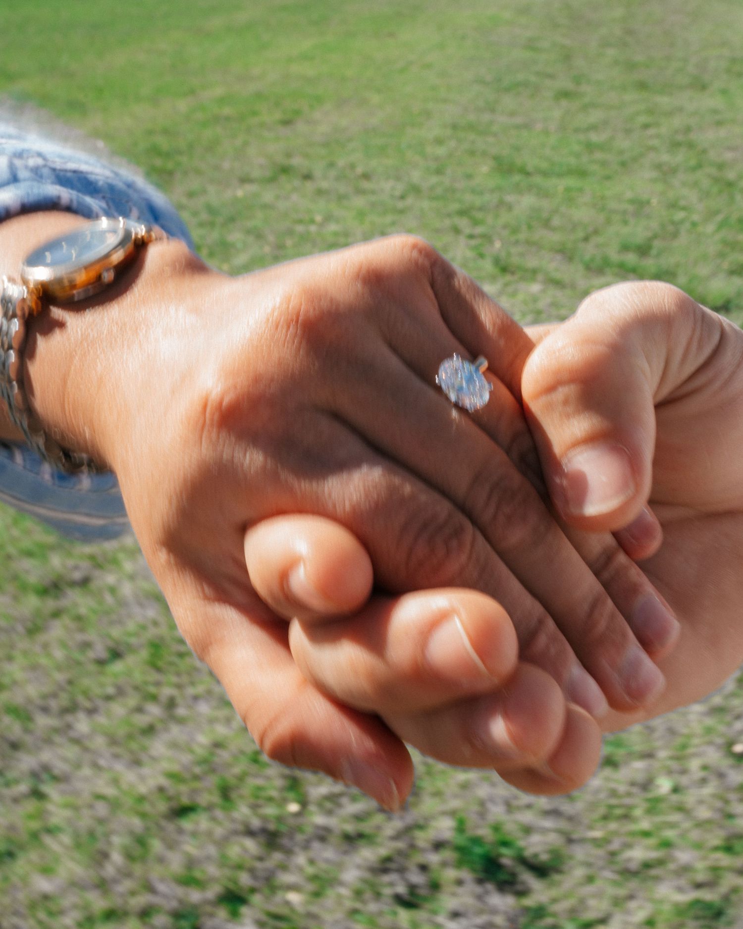 Close-up of hands holding each other with a sparkling diamond engagement ring on display outdoors on a grassy field.