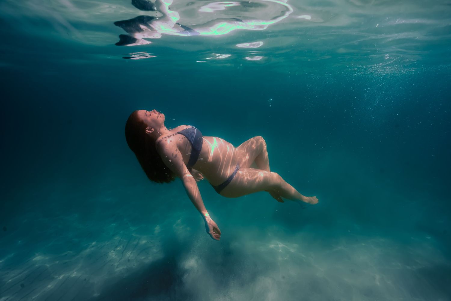 Underwater view of a person swimming gracefully in turquoise blue water with sunlight filtering through the surface.