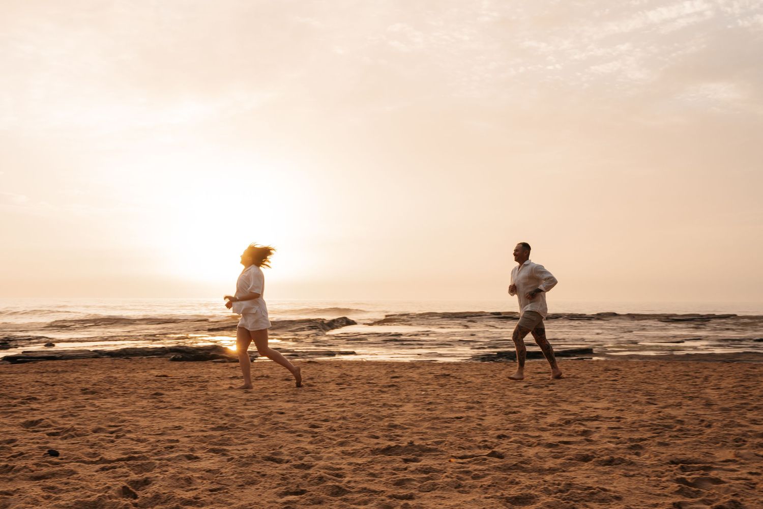 Two people jogging along a sandy beach at sunset with ocean waves in the background.