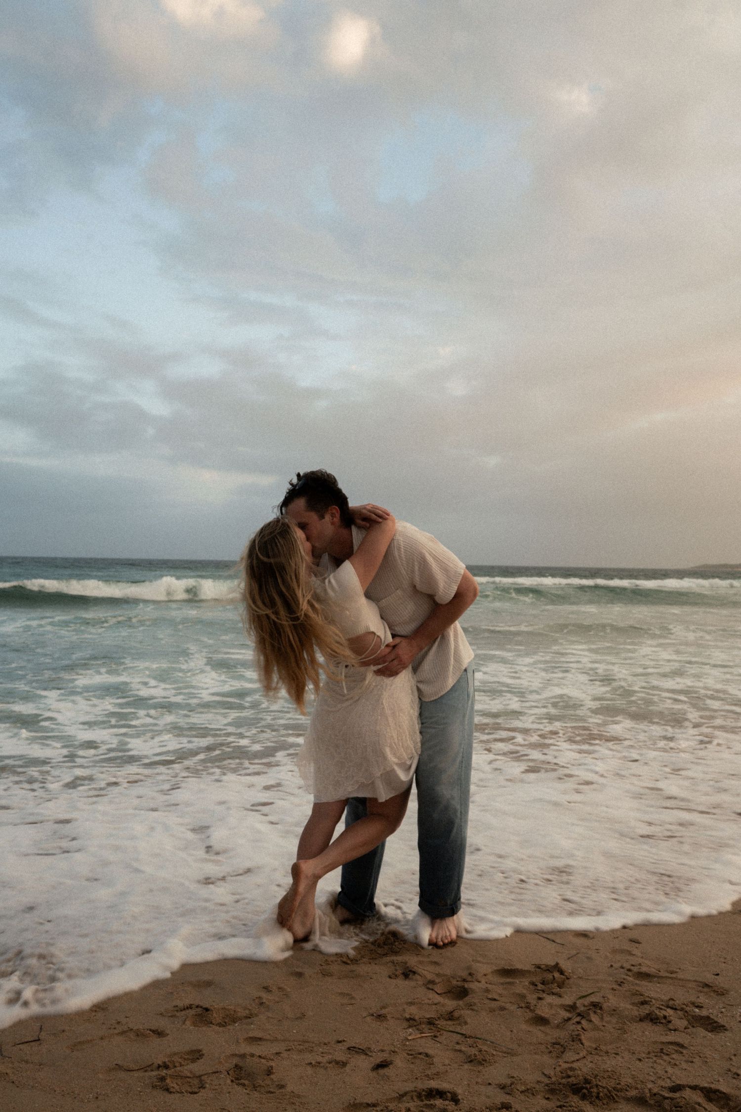 A couple dances playfully together on a sandy beach with ocean waves and cloudy skies in the background.