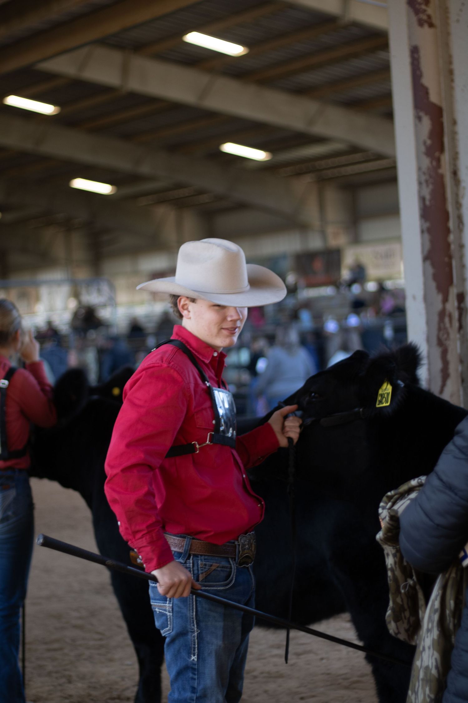 A young person in a red shirt and cowboy hat stands next to a black horse at an indoor equestrian event.