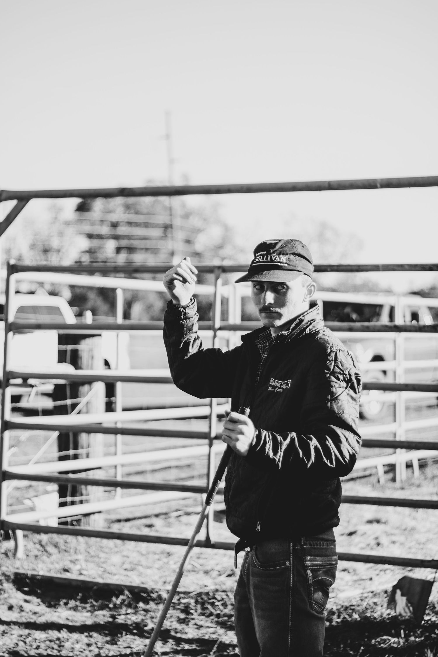A toddler in a winter coat leans against a wooden fence rail at a farm on a sunny day.