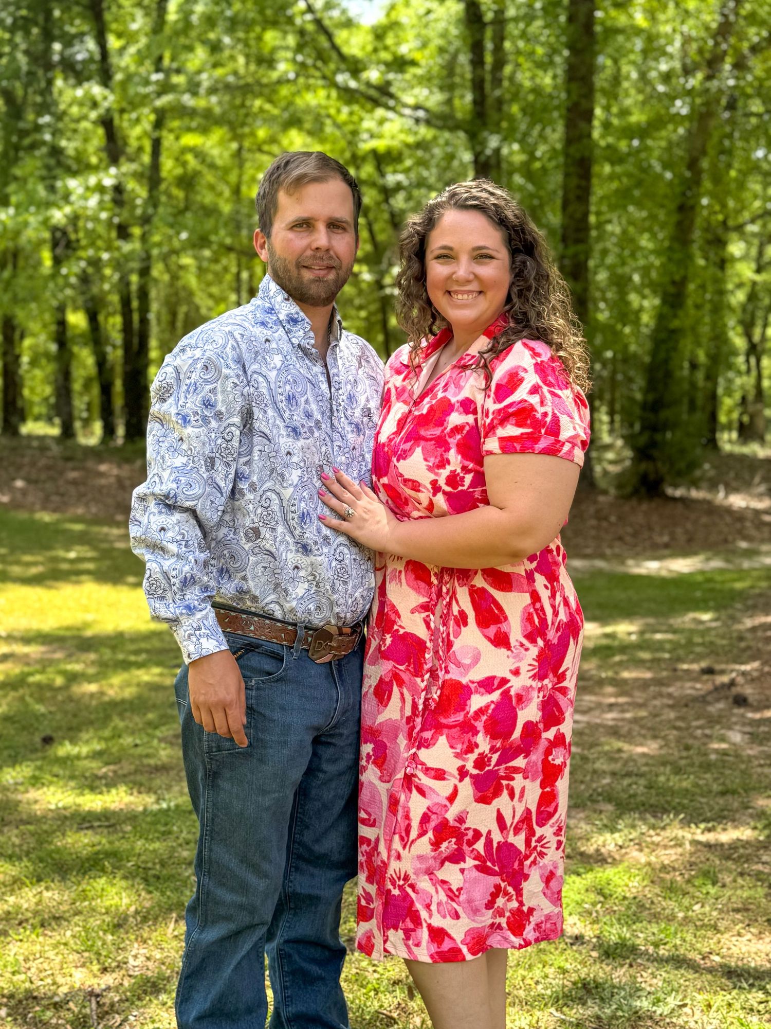 A couple in coordinating red floral and blue outfits pose together for outdoor photos in a sunlit wooded setting.