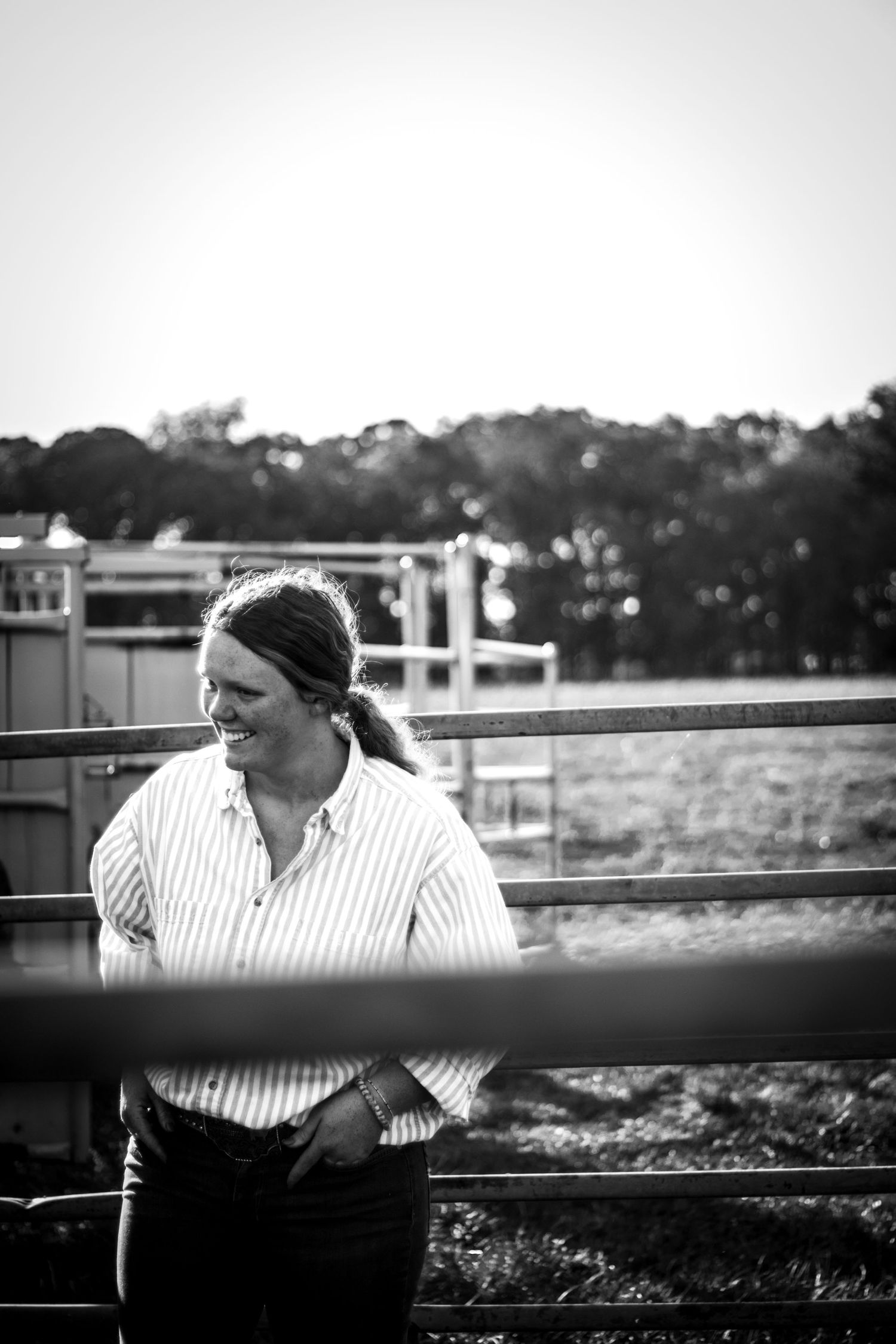 Black and white portrait of someone in a striped shirt leaning against a fence railing at sunset.
