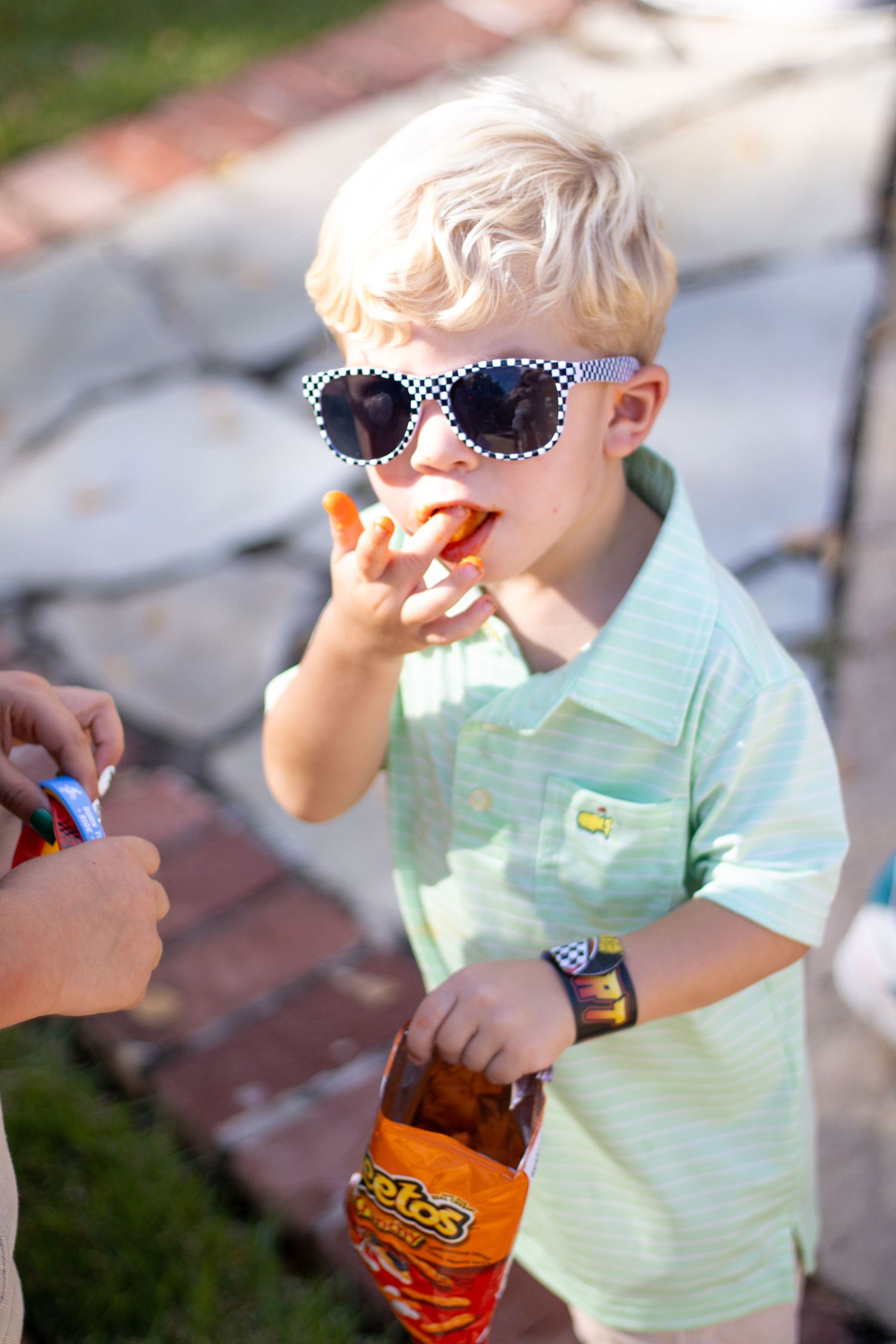 A child in sunglasses and mint green shirt eating snacks outdoors.
